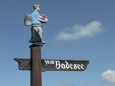 Wegweiser aus Holz mit Figur in Freizeitkleidung mit Rucksack und einem Wasserball in der Hand Wegweiser aus Holz mit Figur in Freizeitkleidung mit Rucksack und einem Wasserball in der Hand